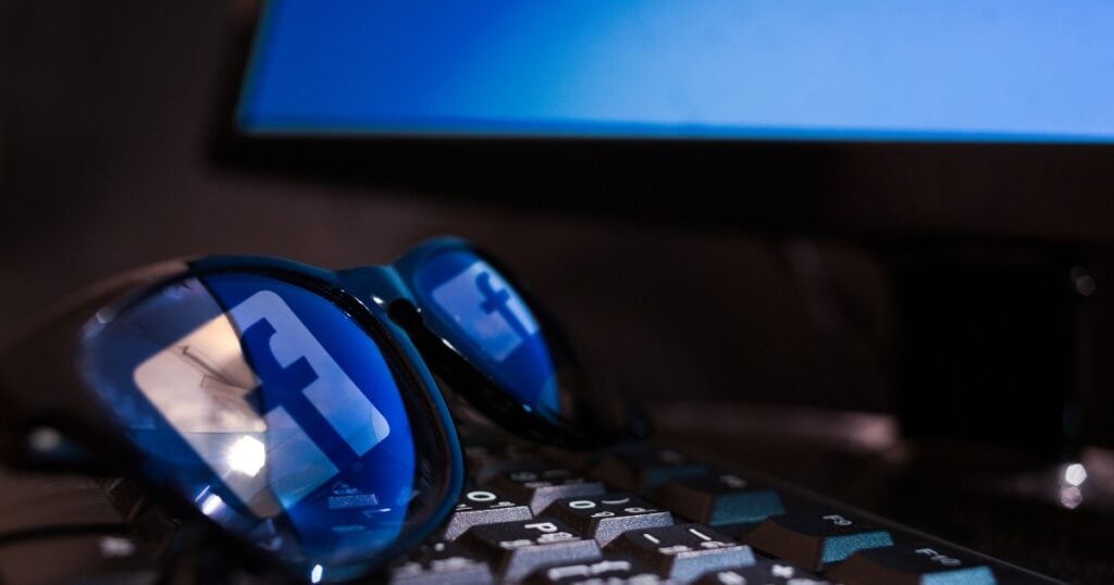 Reflection of a Facebook logo from a computer screen on sunglasses positioned above a keyboard.
