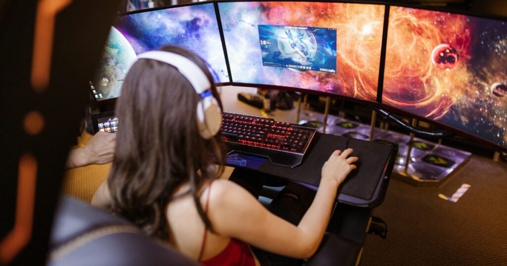 A girl with white headphones plays a video game on a desktop computer with three screens.