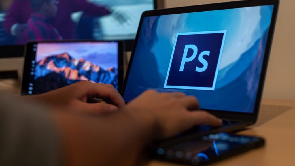 person typing in his laptop while the software adobe photoshop is opened in the screen